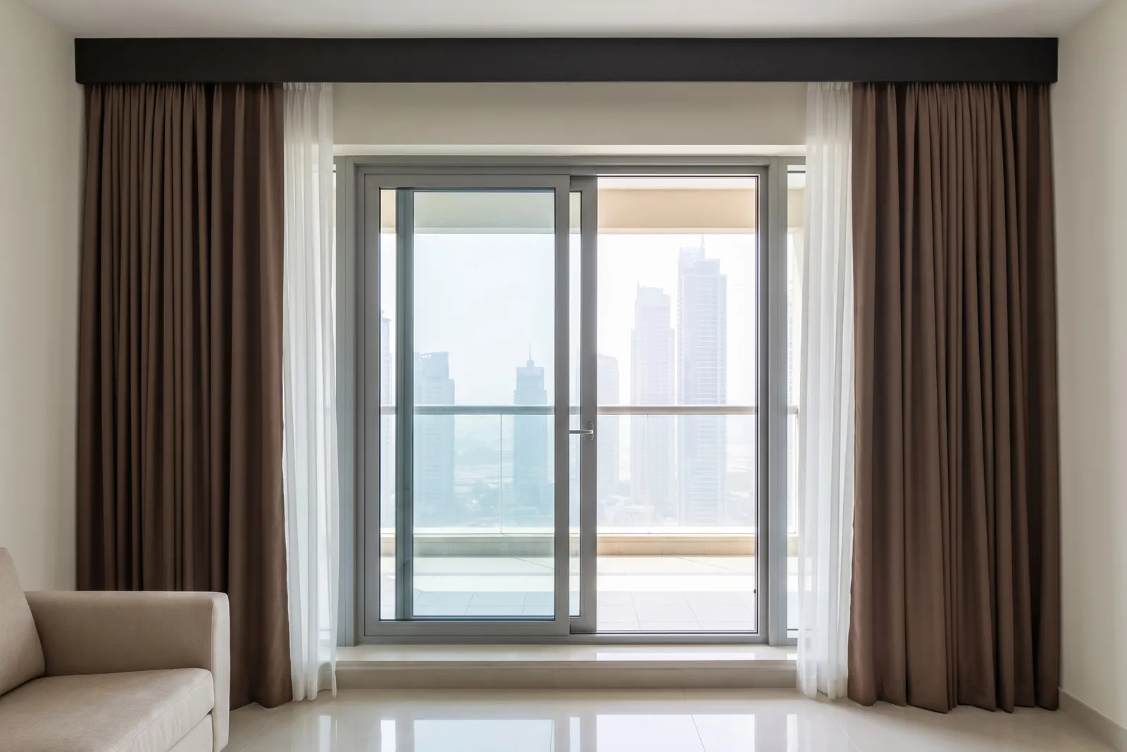 Mocha brown lined curtains and white sheers drawn open at both sides of a sliding balcony door, towers beyond, a sofa arm at the left