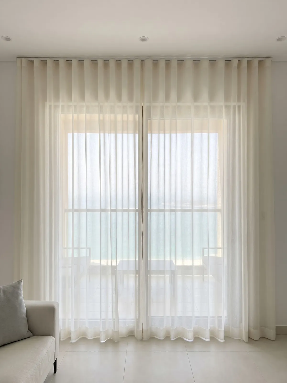 Ivory sheer pair closed across a sliding balcony door, the balcony chairs and the sea soft behind it, a pale sofa arm at the lower left