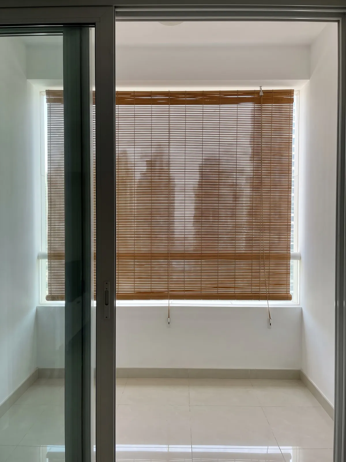 Caramel roll-up bamboo blind lowered across a covered balcony, seen through an open sliding door, towers soft through the weave