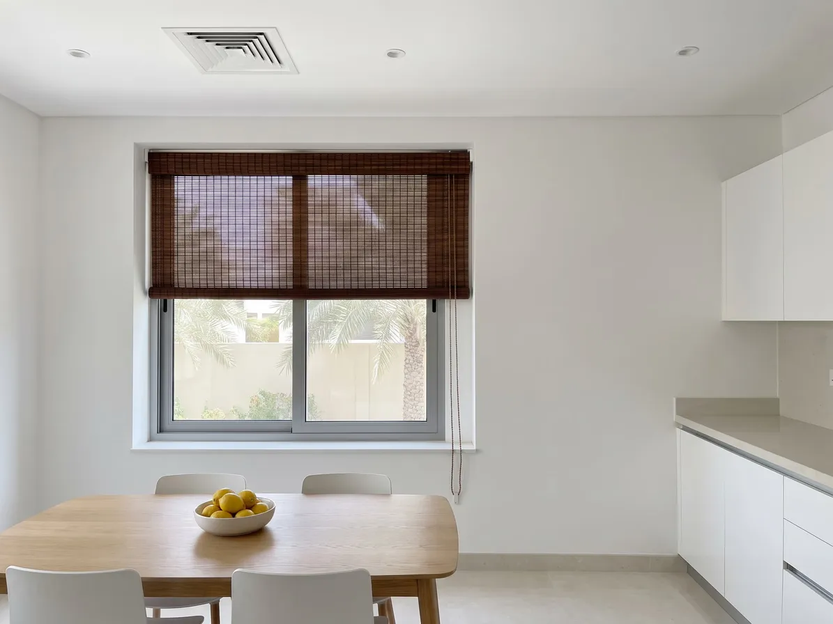 Dark toasted bamboo blind half lowered on a kitchen-diner window behind a pale oak table with a bowl of lemons, palms below
