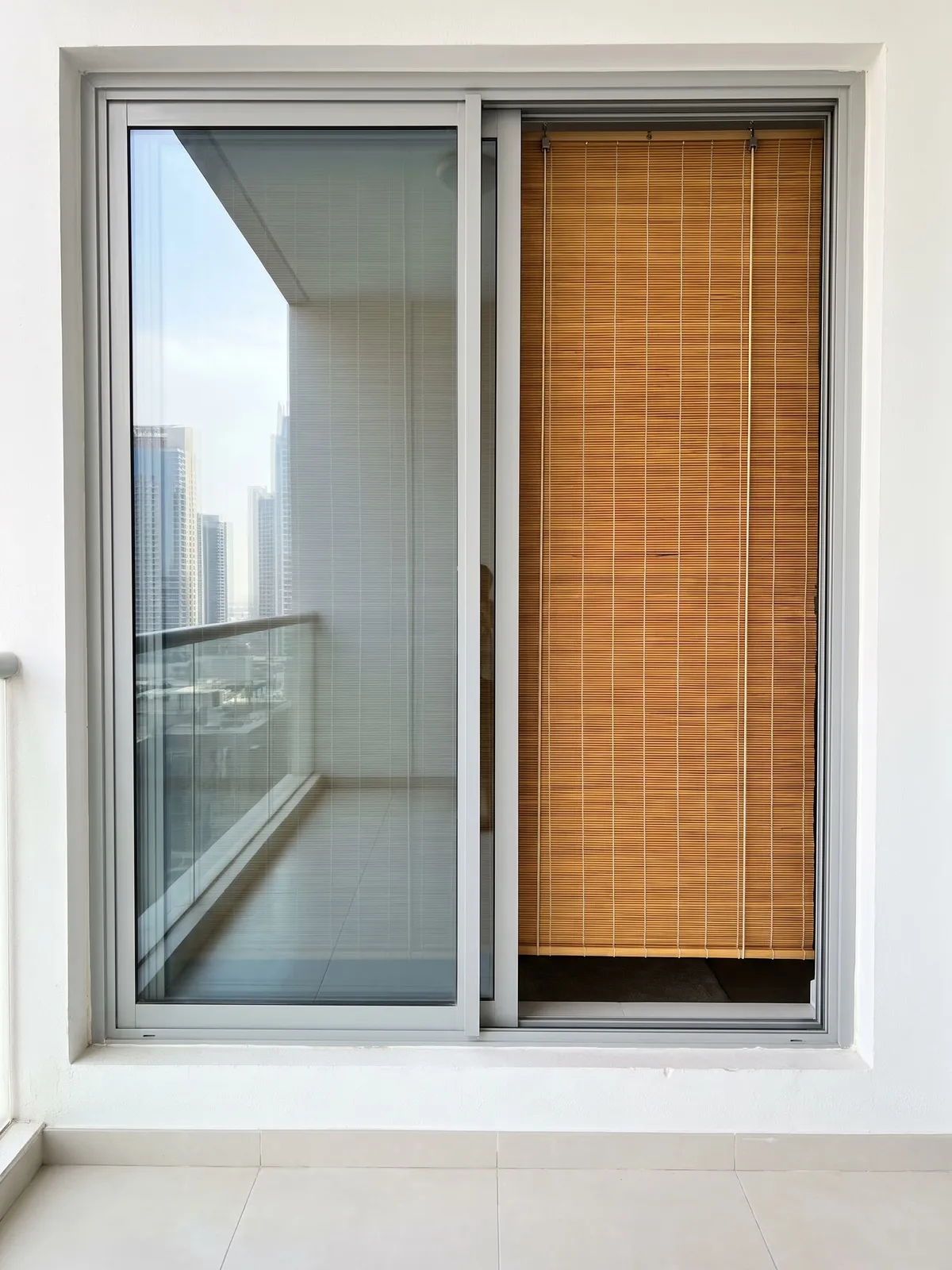 Back of an amber bamboo blind lowered behind one pane of a sliding door, seen from the balcony, towers in the other pane