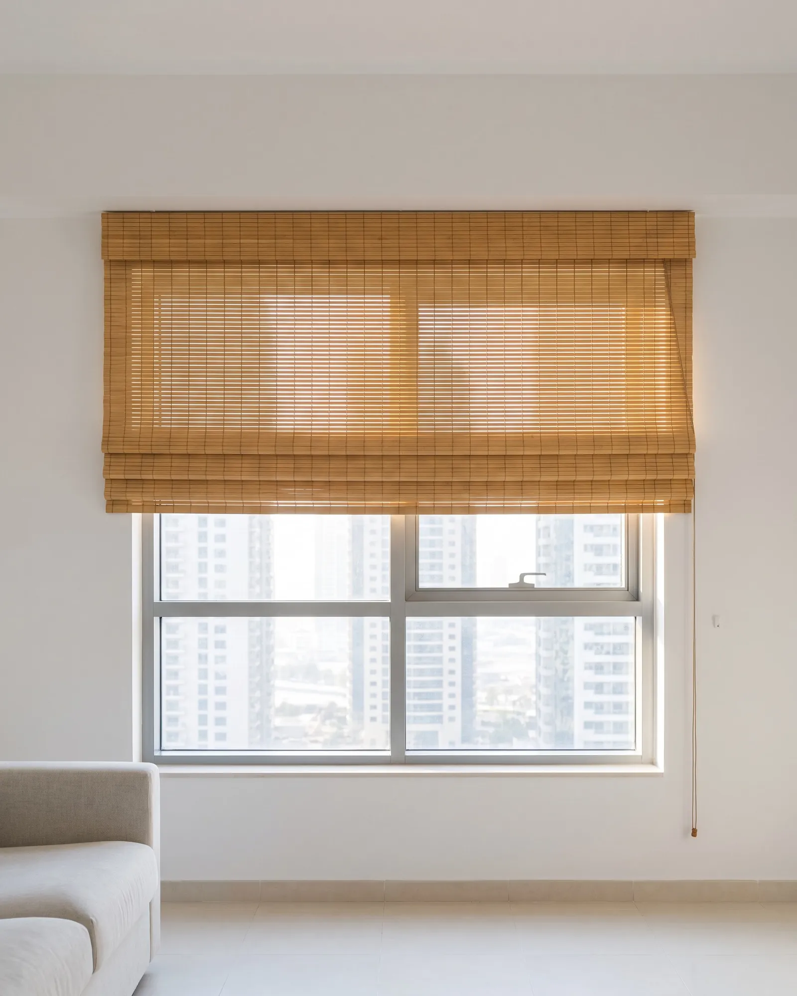 Honey bamboo blind lowered over the top half of a living-room window, afternoon light through its weave, towers below, a sofa at the left
