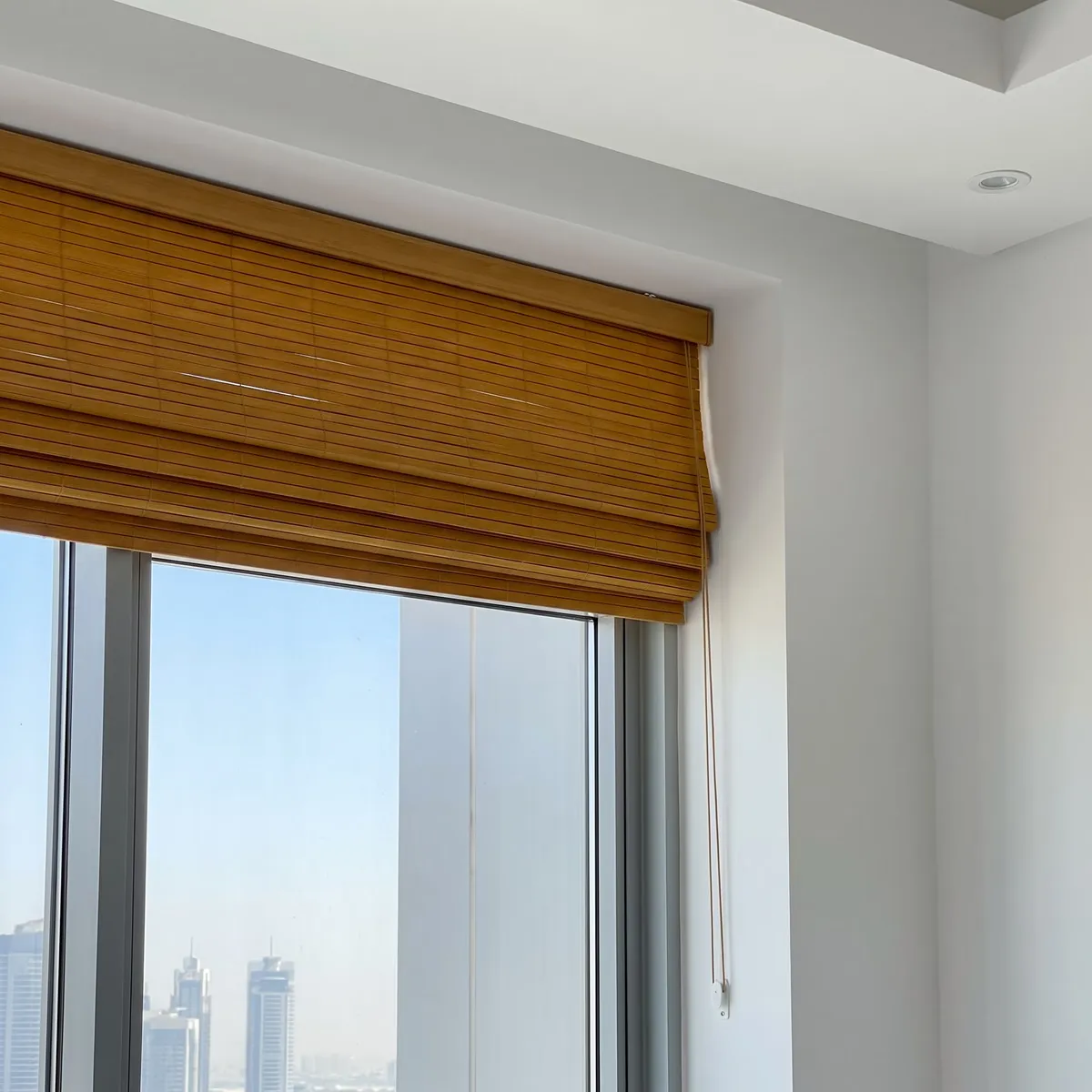 Golden honey Roman-fold bamboo blind raised at the top of a tall window, its pull cord running down the right side, towers below