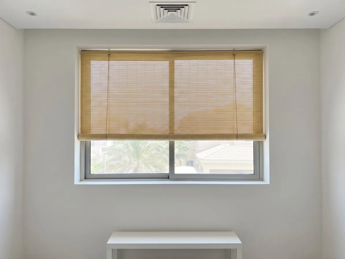Pale straw roll-up bamboo blind lowered over the top two thirds of a window, its cord loops down the front, a white console below