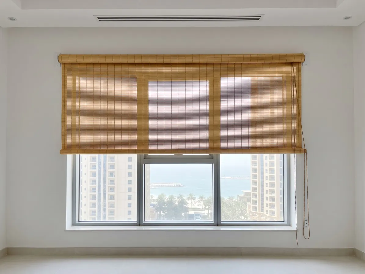 Light honey bamboo roller blind lowered over the top half of a wide window, its roll on the wall above, the sea and towers below