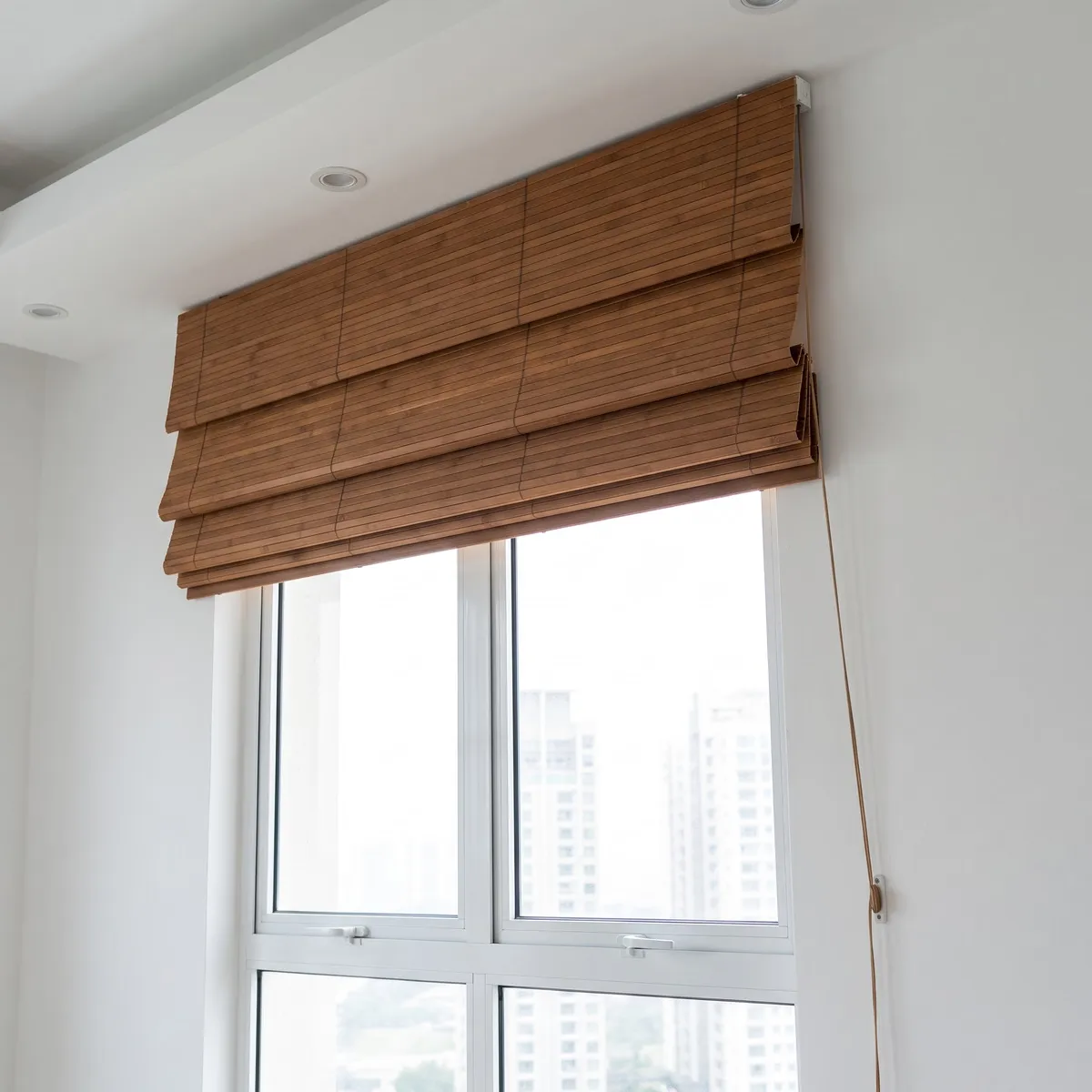 Toasted brown Roman-fold bamboo blind raised at the top of a tall window, seen from the side with its folds stacked, towers below