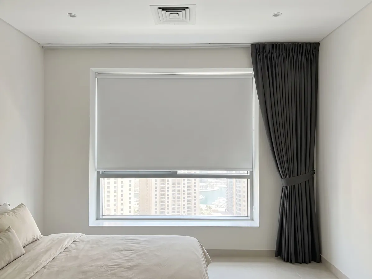 White roller blind lowered inside a window recess, a charcoal pleated curtain tied back beside it, towers in the glass below the blind