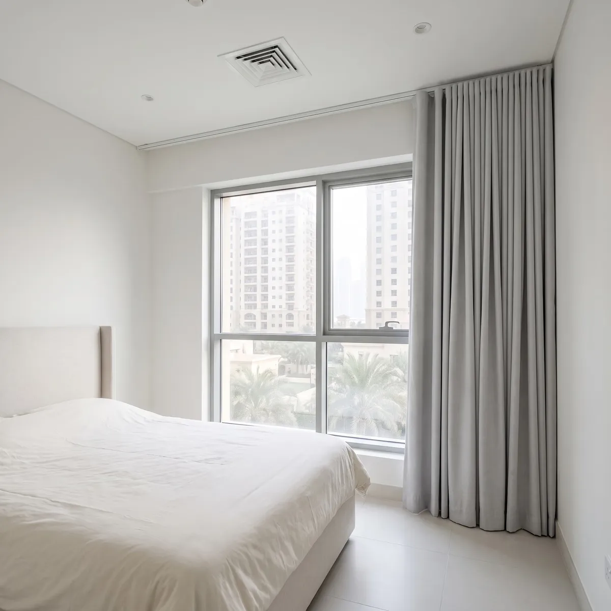 Light grey wave curtain gathered at the end of a ceiling track away from the bed, a window with towers and palms beside it