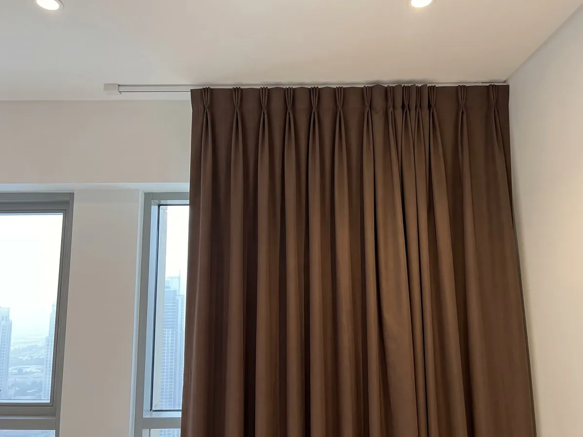 Top of a closed mocha-brown pleated curtain under a white ceiling track, small pleats in a row, a window with towers at the left