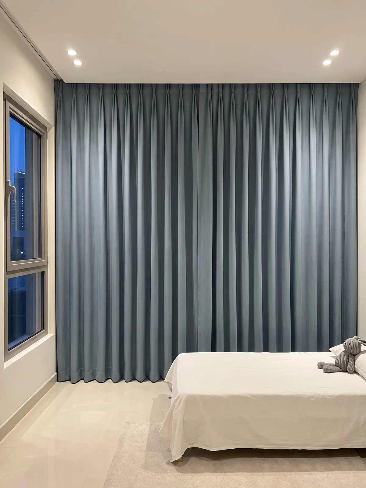 Dusty-blue pleated curtains closed ceiling to floor in a child's room at dusk, a low bed with a knitted rabbit and a window with a blue sky