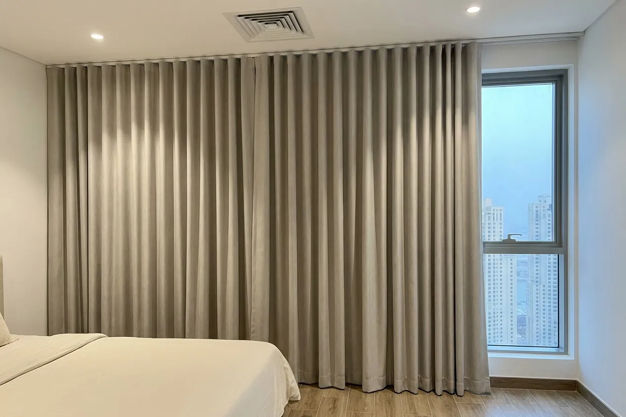 Warm grey-beige wave curtains closed across a bedroom wall, a tall narrow window with towers in blue haze beside them, an ivory bed at lower left