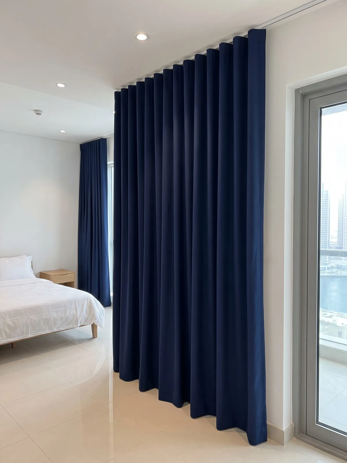 Closed navy wave curtain seen from the side on a ceiling track, its hem at the floor, a glass balcony door at the right and a bed beyond