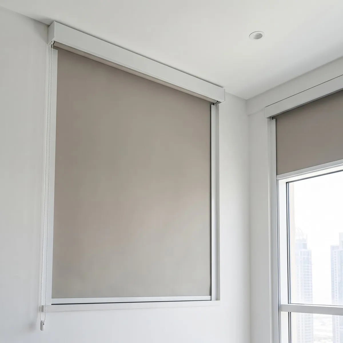 Greige blackout roller blind lowered under a white cassette, seen from below at an angle, a second window with its own blind at the right