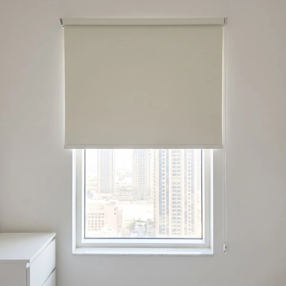 Ivory blackout roller blind lowered halfway down a window, its chain held straight by a small clip at the right, towers below it