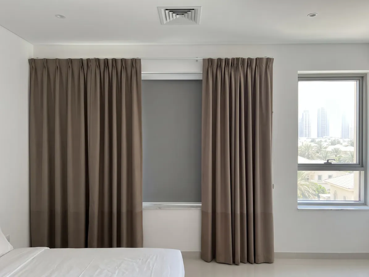 Taupe curtains open at both sides of a window with a grey blackout roller lowered between them, a bed in front, towers at the right