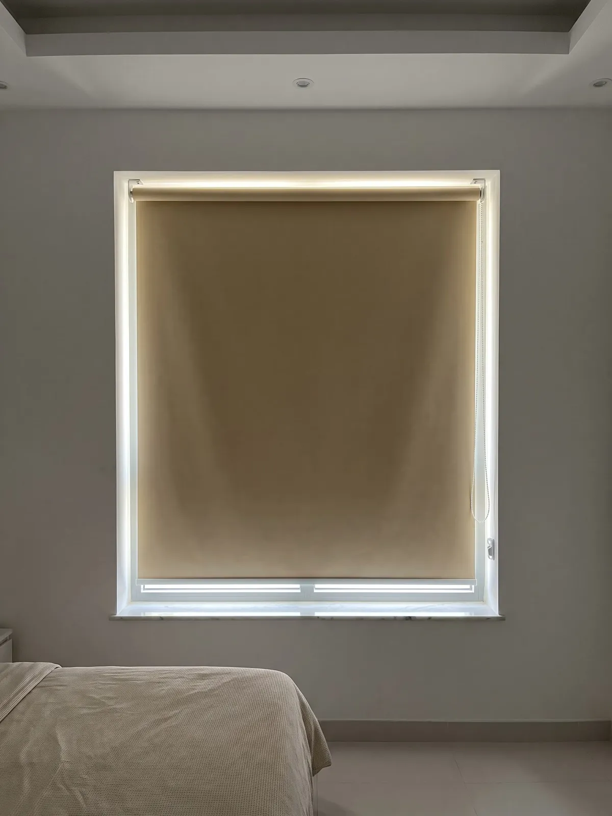 Sand blackout roller blind lowered in a window recess of a dim bedroom, daylight glowing along its edges, a bed at the lower left