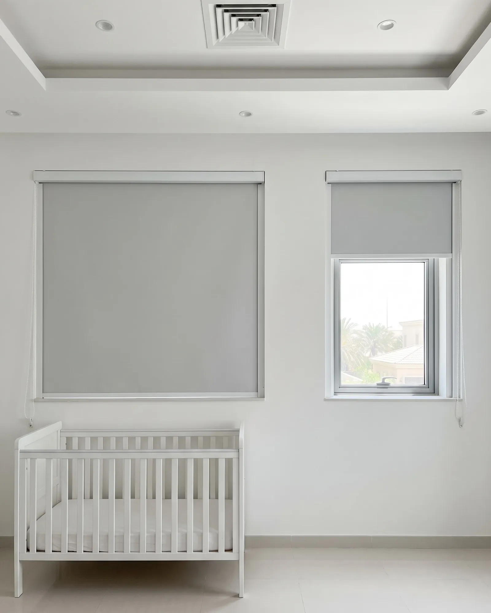 Light grey blackout roller blinds in a nursery, one lowered on the large window, one raised on a small window, a white cot below
