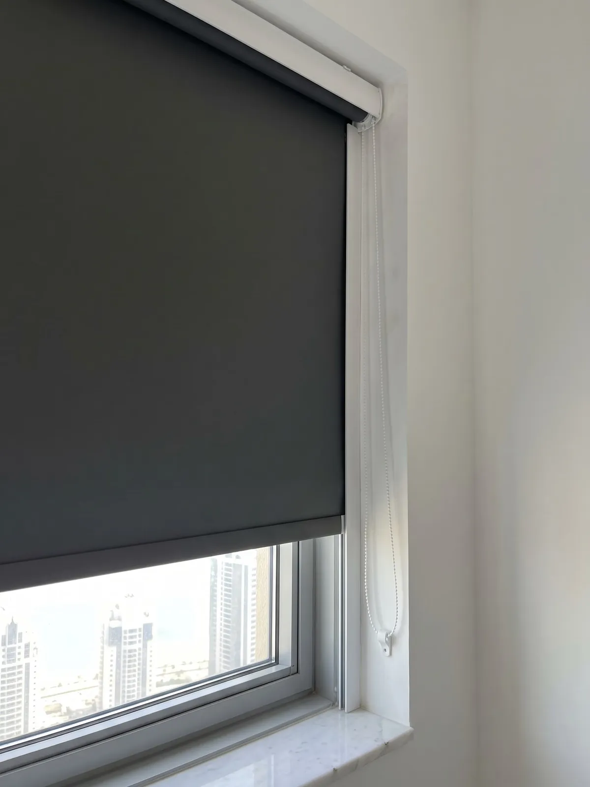 Charcoal blackout roller blind lowered most of the way down a window, a white chain at its right edge, towers showing below its bar