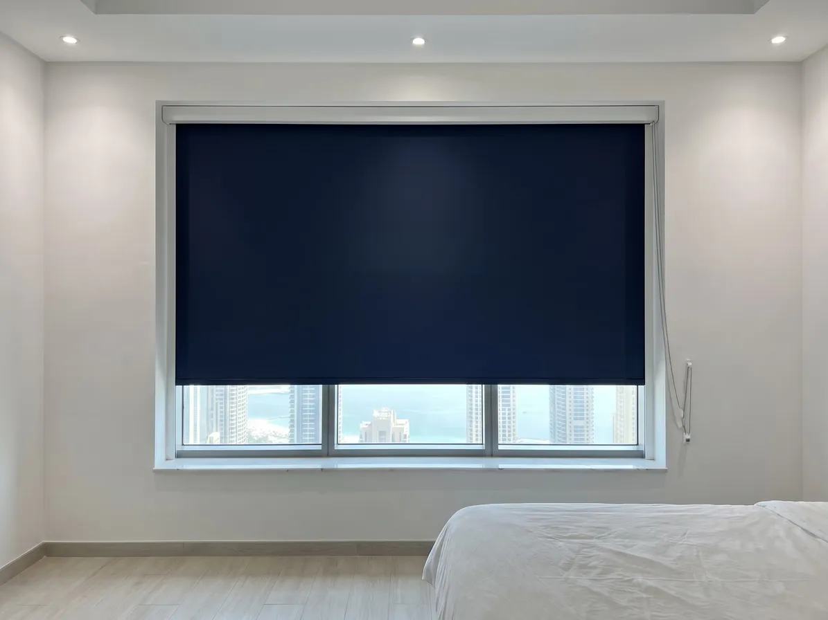 Navy blackout roller blind lowered most of the way on a wide bedroom window, the sea and towers below its bar, a bed at the right