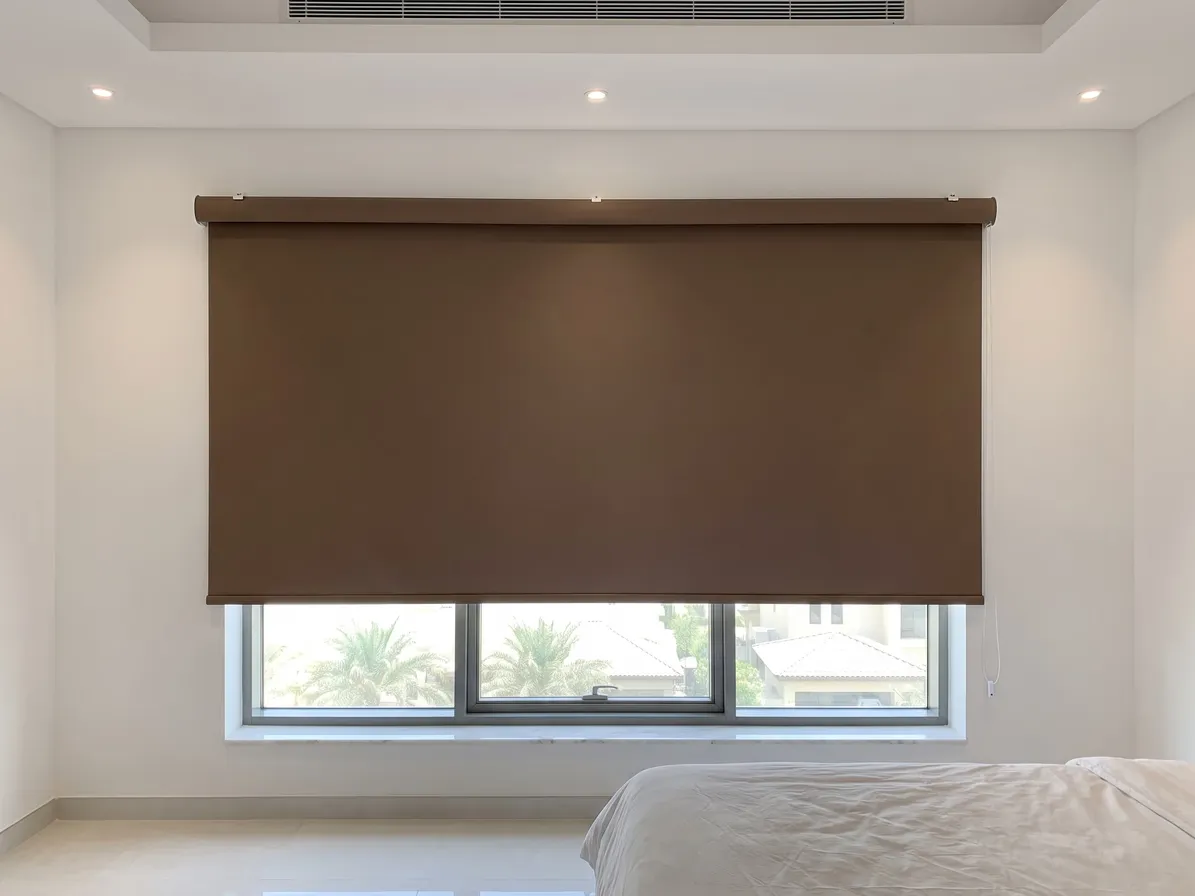 Mocha brown blackout roller blind lowered over a large bedroom window, running past its frame, palms and villas below, a bed at the right