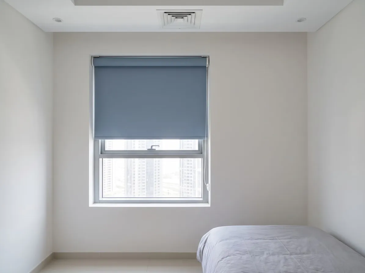 Dusty blue blackout roller blind half lowered inside a small bedroom window recess, towers below it, the foot of a bed at the right