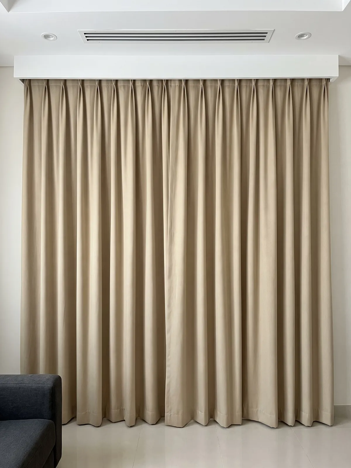 Sand-beige blackout curtains with pinch pleats closed under a white box, one even colour, a dark sofa arm at lower left