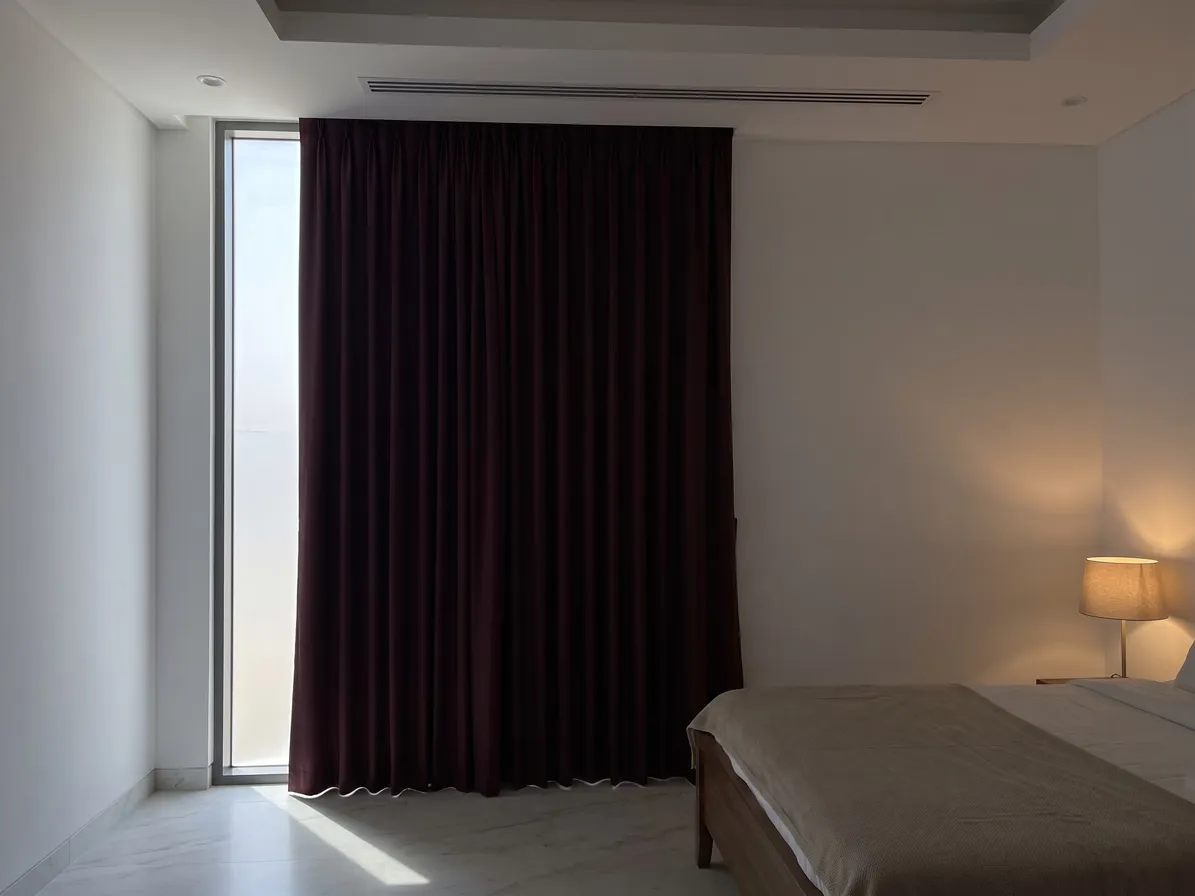 Dark burgundy blackout curtain closed in a bedroom, one tall strip of bright glass beside its left edge, a lamp lit at right