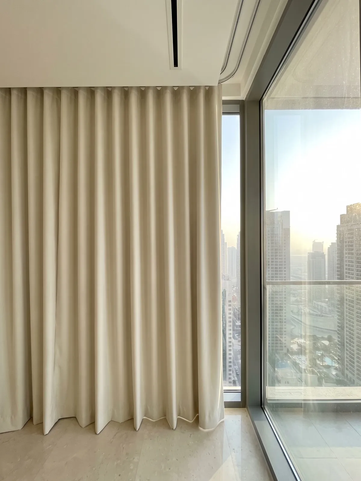 Cream blackout curtain in a wave heading closed beside a tall corner window, towers in hazy sun beyond the glass