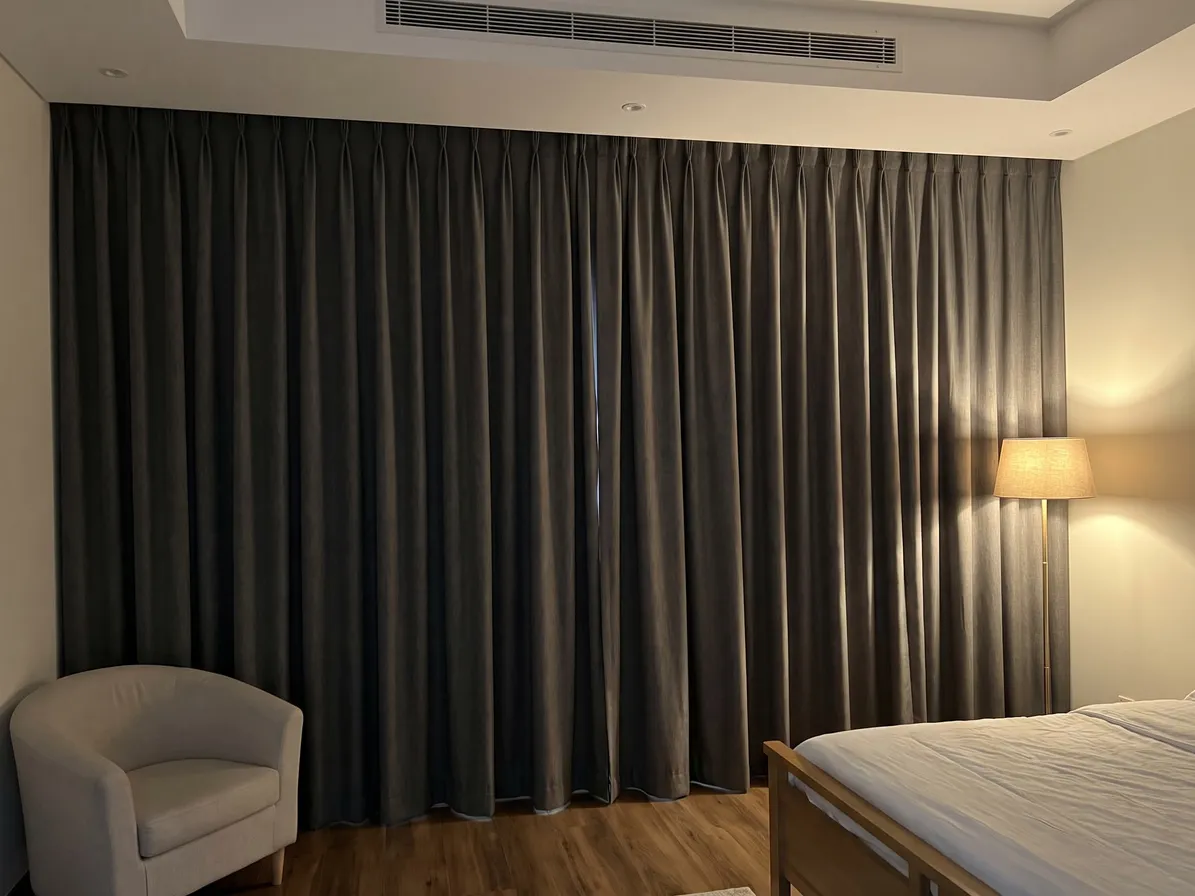 Dark grey-brown pinch-pleat curtains closed across a bedroom wall, a tub chair, a lit floor lamp and the foot of a bed