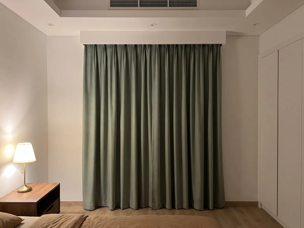 Sage-green pinch-pleat curtains closed under a white box, a lit lamp on a wooden nightstand, a white wardrobe at right