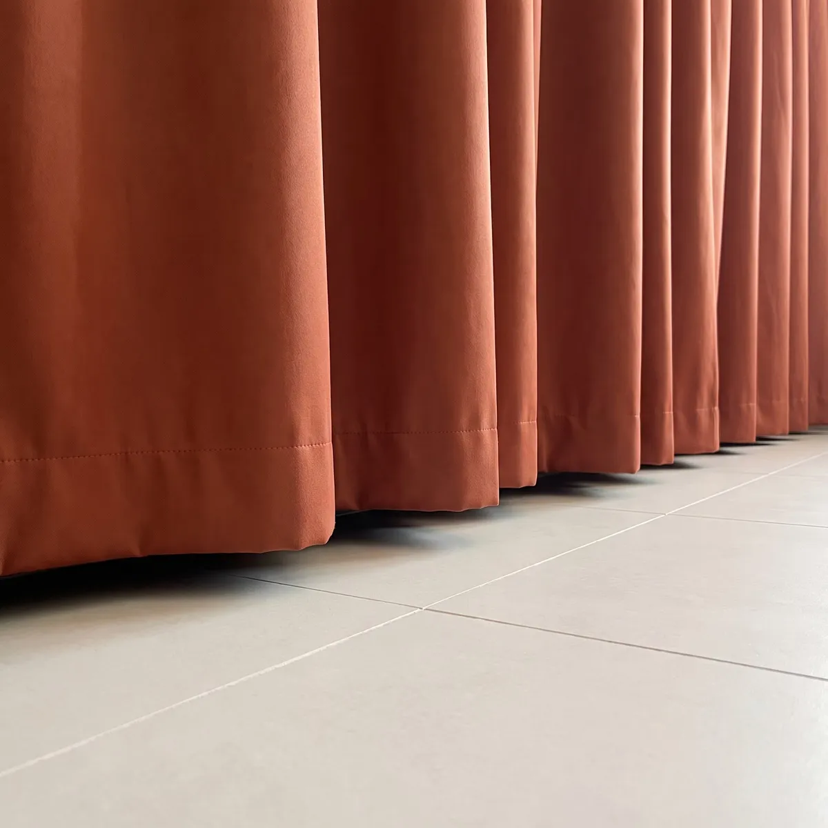 Bottom of a closed terracotta curtain, soft straight folds falling into a stitched hem just above a pale tile floor