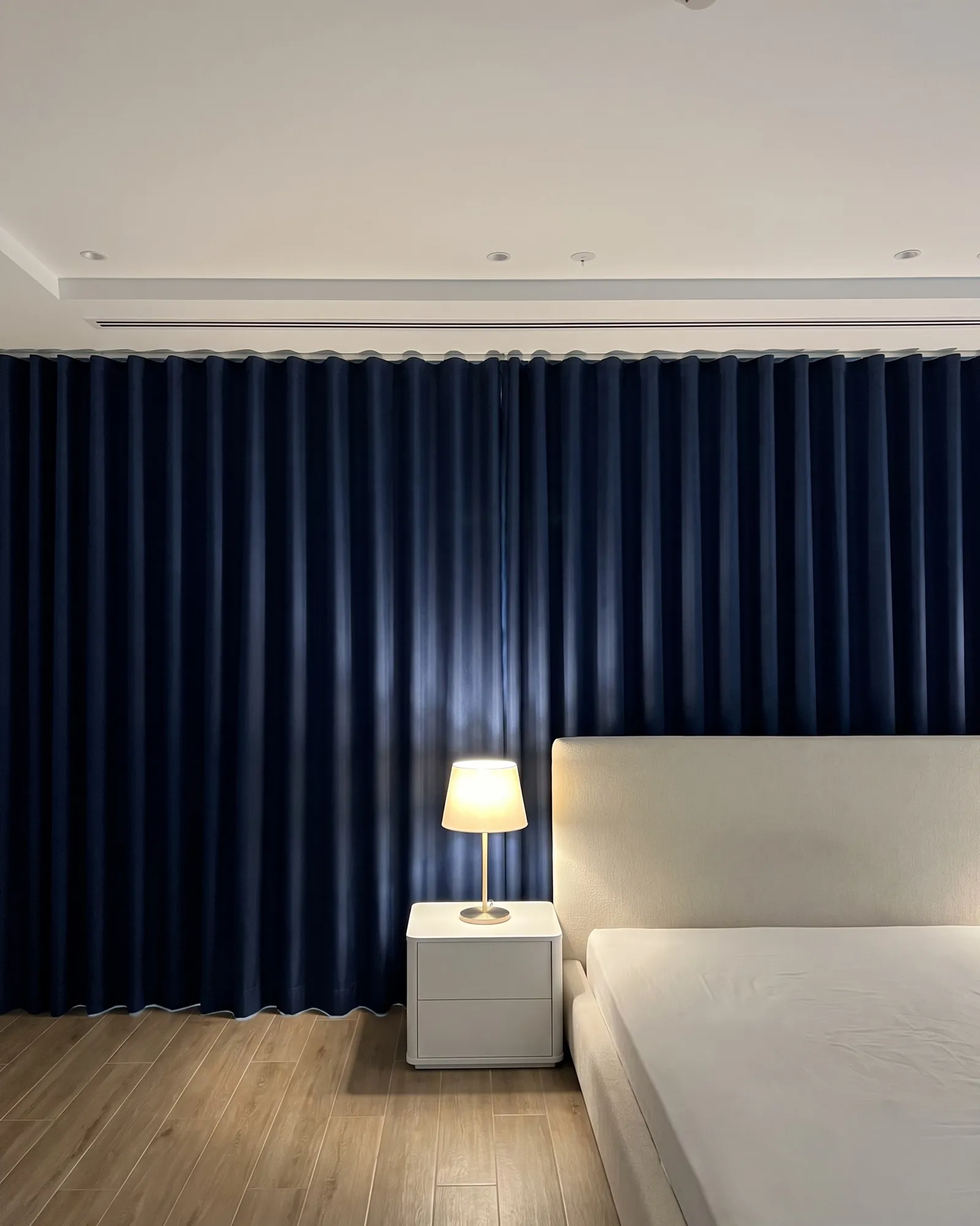 Deep navy blackout curtains in a wave heading closed across a bedroom wall, a lit bedside lamp and an ivory headboard in front