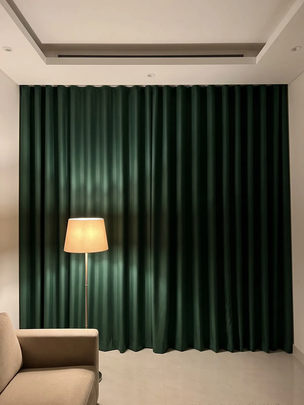 Dark green blackout curtains in a wave heading closed from ceiling to floor, a lit floor lamp and a beige sofa arm in front