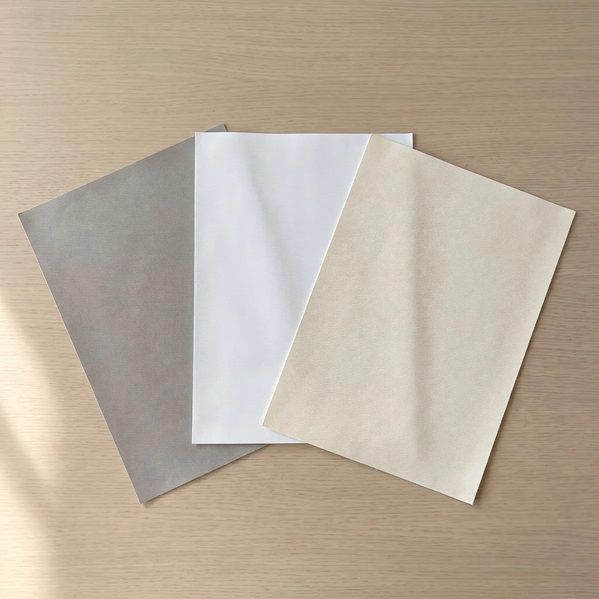 Three flat fabric samples, grey, white and cream, fanned out on a pale oak table in window light