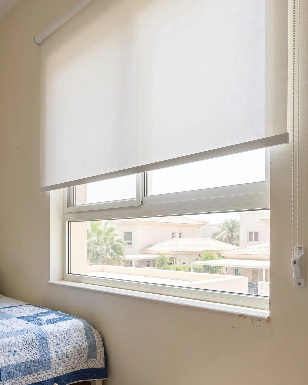 White roller blind in a child's bedroom, its chain held taut beside the window, a patterned quilt below