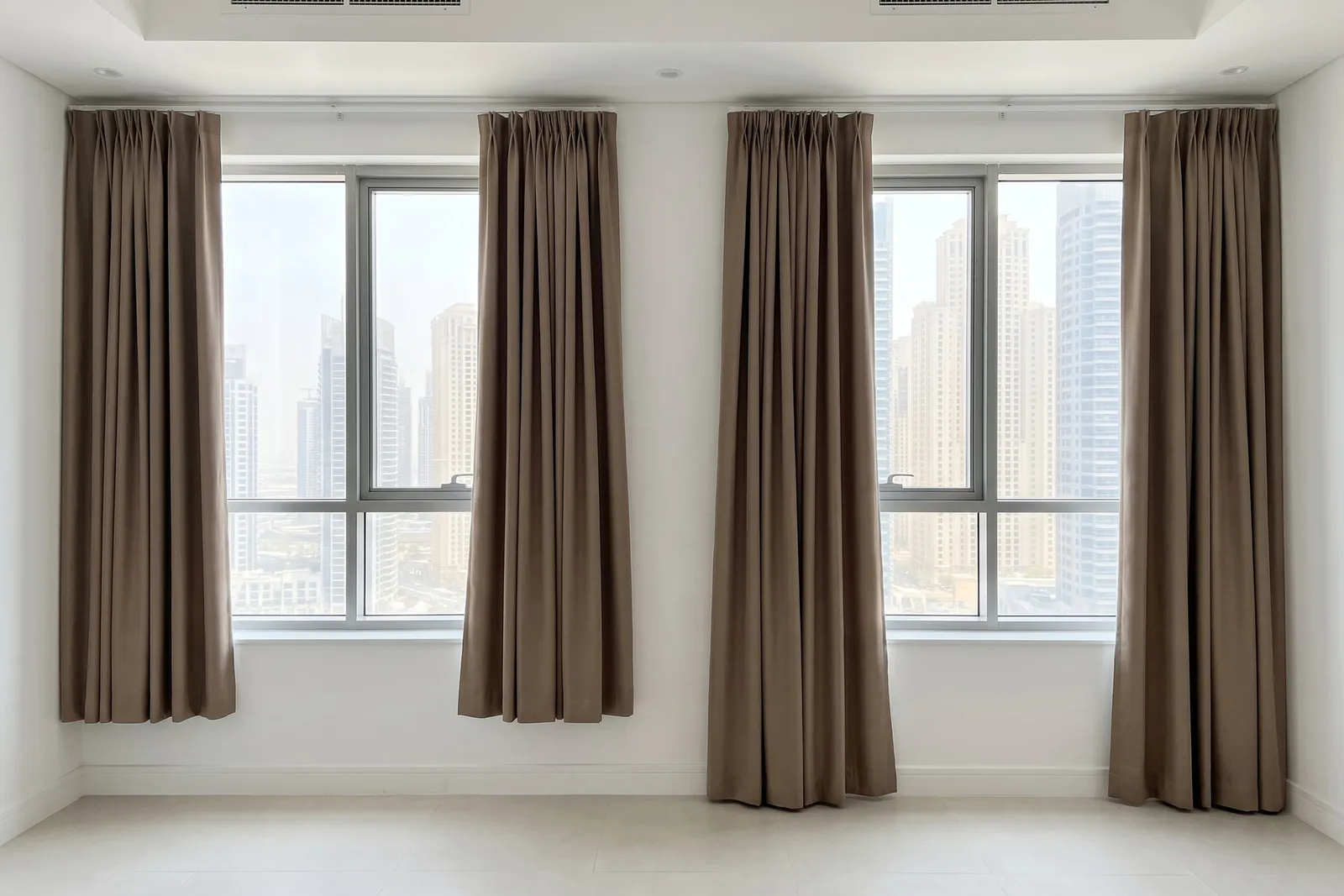 Taupe curtain pairs on two matching windows, the left pair stopping well short of the floor and the right pair reaching it, towers outside