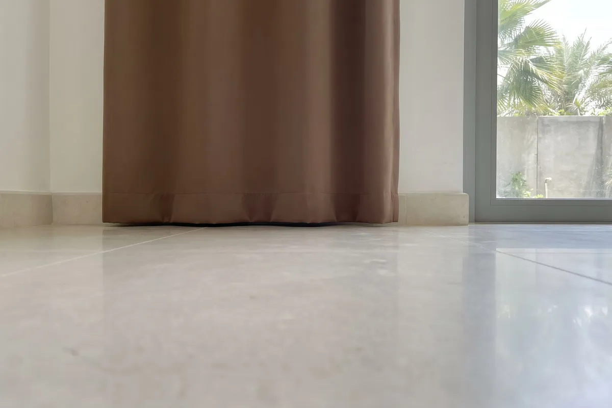 The hem of a mocha curtain just meeting a pale stone floor, seen at floor level, a glass door with palms at the right