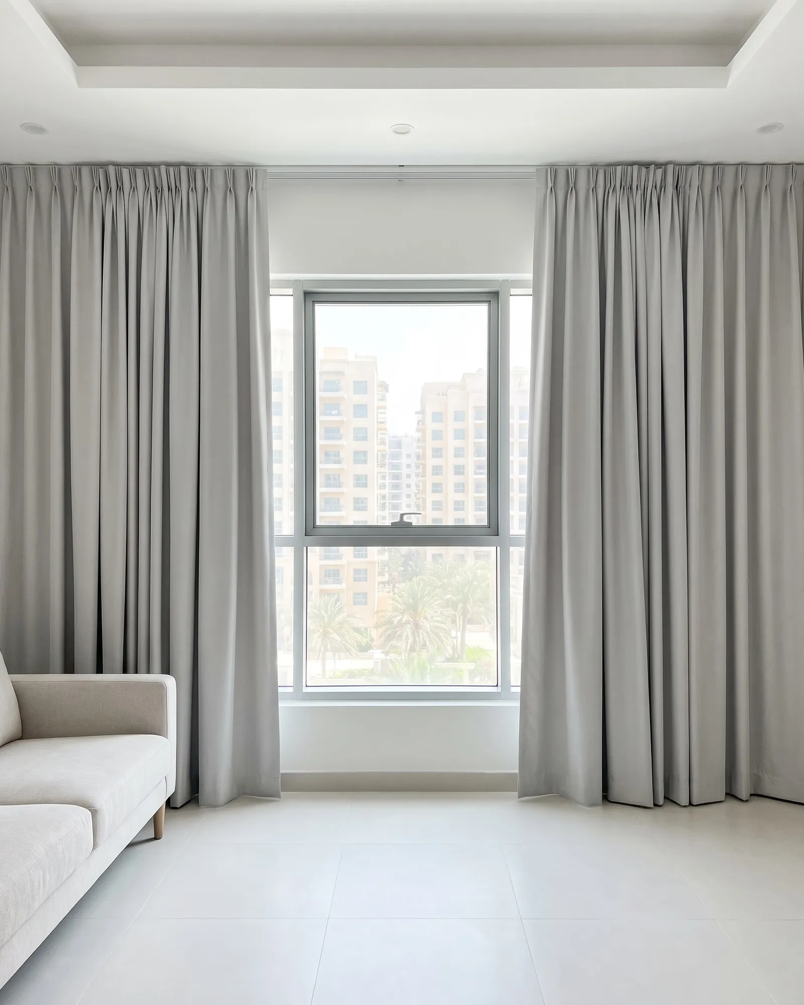 Light grey lined curtains drawn open at both sides of a living-room window, apartment blocks and palms outside, a pale sofa at the left