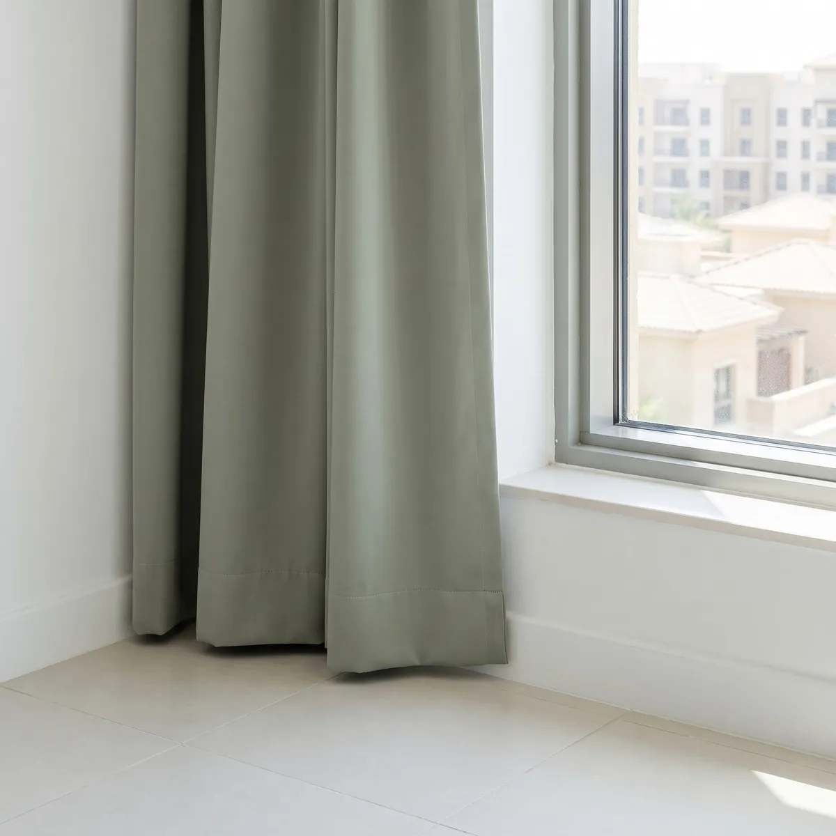 Sage curtain hanging beside a window, its hem just clear of the floor, villa roofs and apartment blocks outside