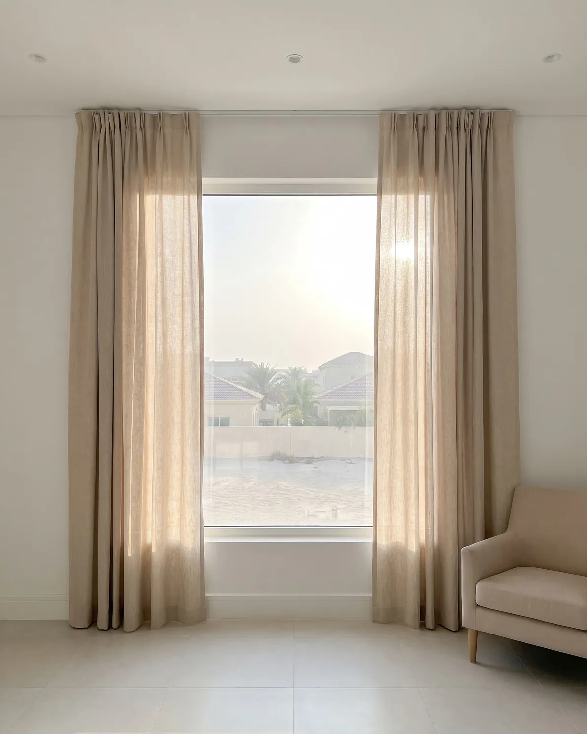 Oatmeal linen curtains drawn open at both sides of a sunny window, light glowing through the cloth, an armchair at the right