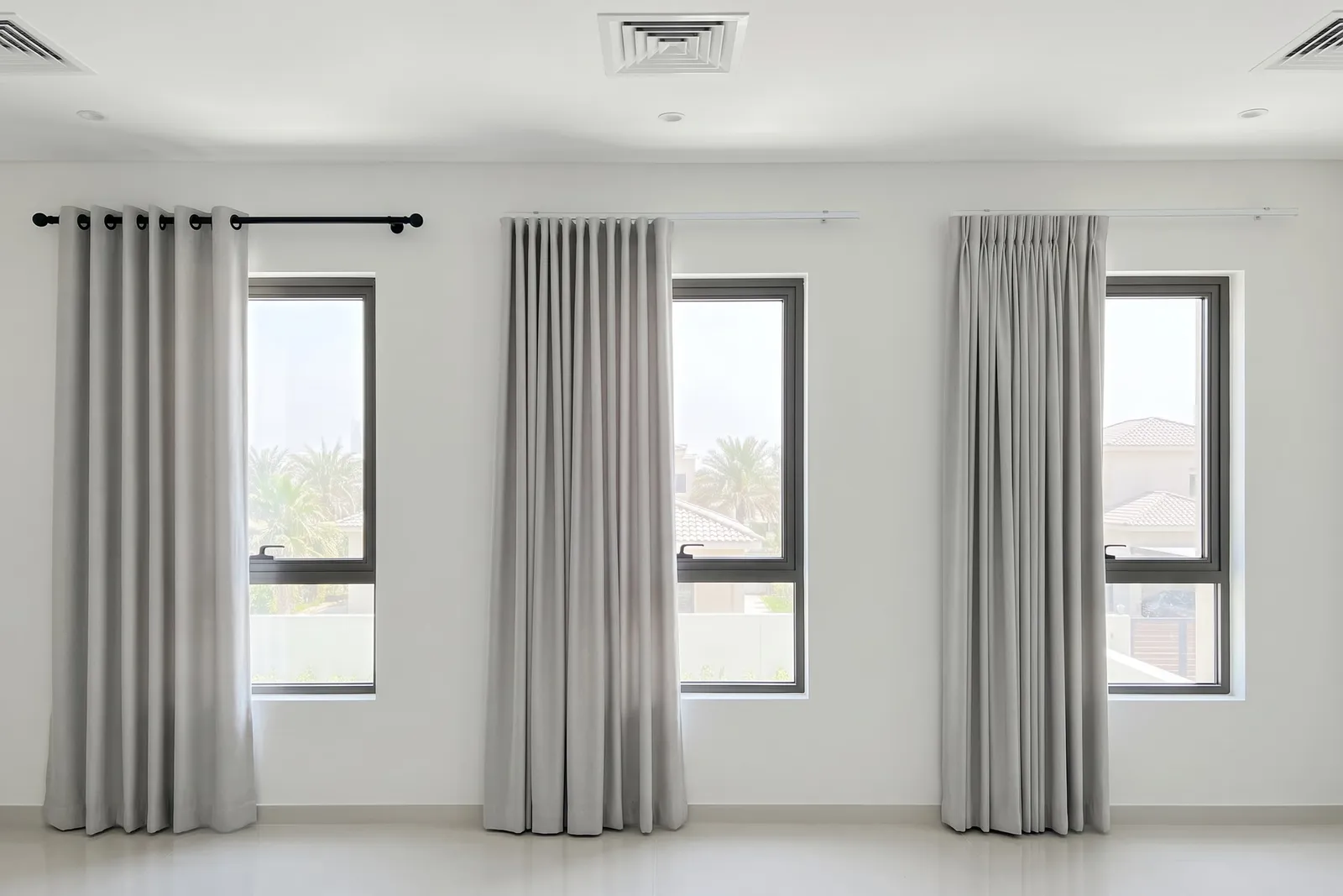 Eyelet, wave and pinch pleat curtains in light grey hung beside three windows of one room, palms and villa roofs through the glass