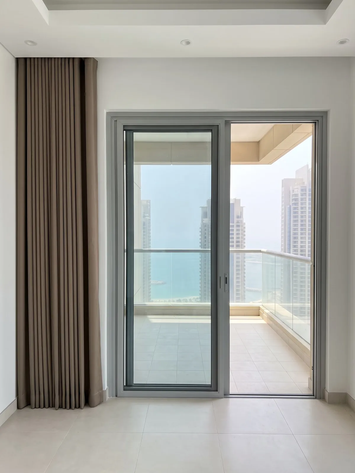 Taupe wave curtain gathered beside an open sliding balcony door, a glass railing with the sea and towers beyond