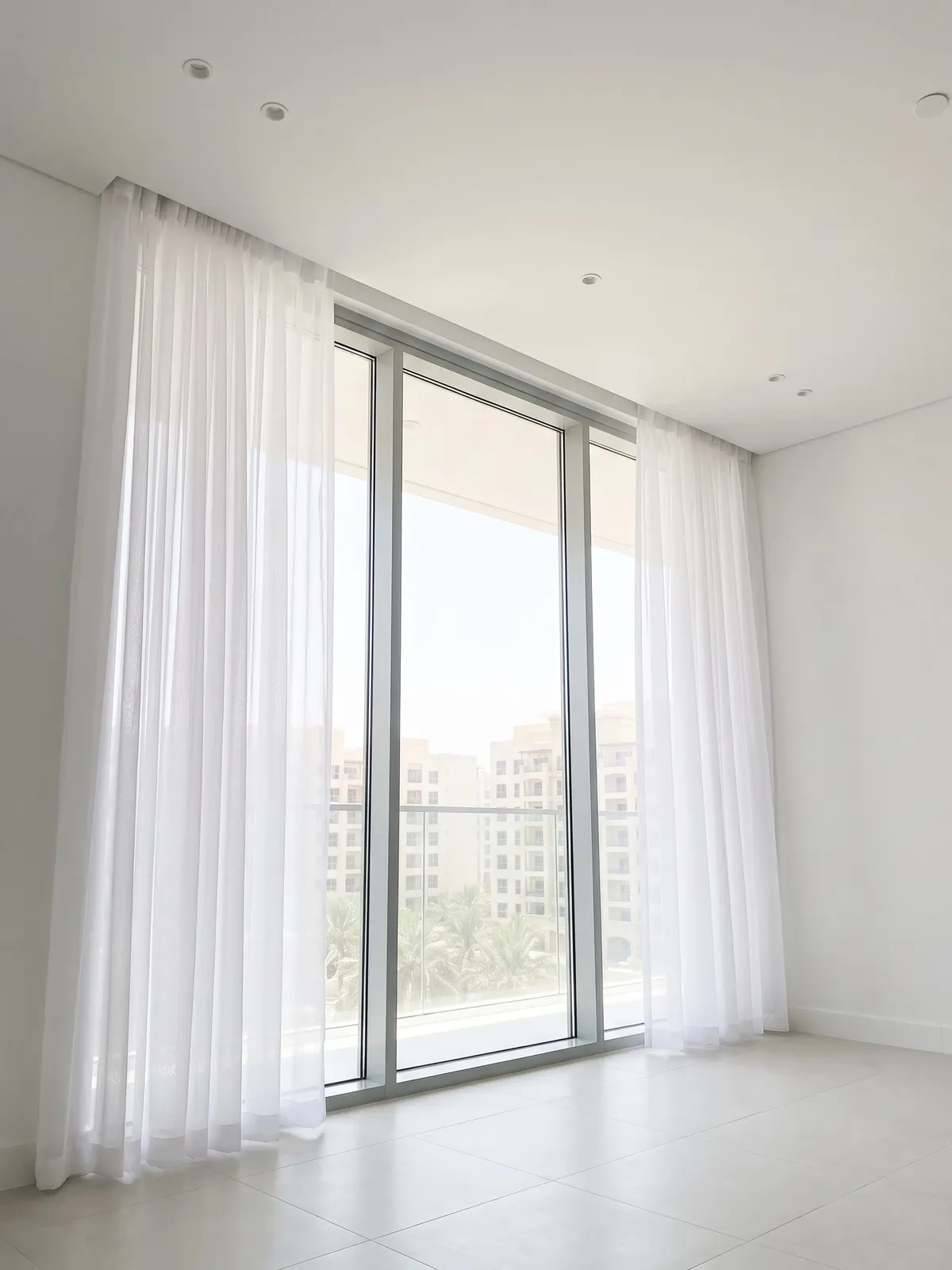 White sheers hanging from the ceiling, drawn open at both sides of floor-to-ceiling glass, a balcony, palms and apartment blocks beyond