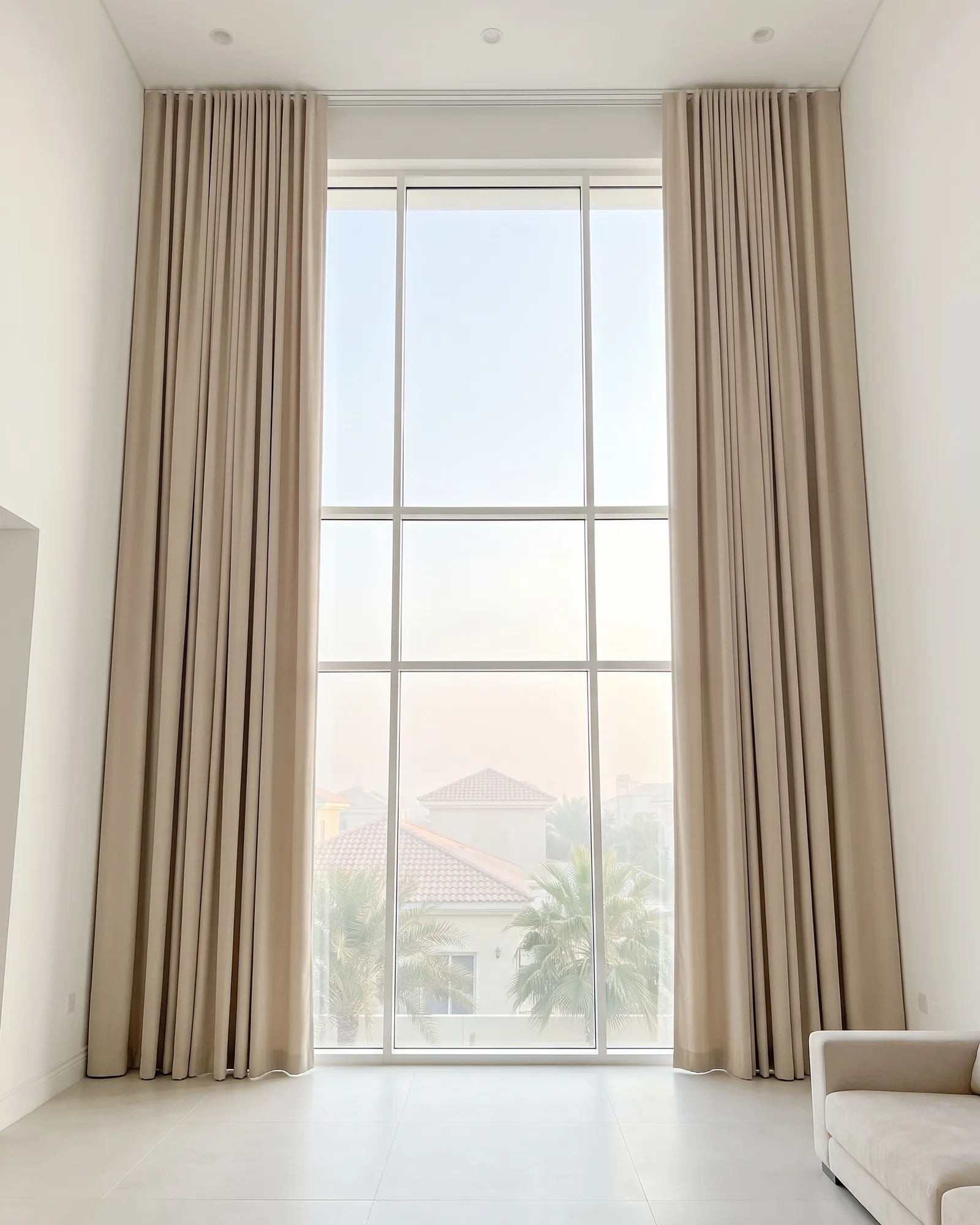 Light beige lined curtains drawn open at both sides of double-height villa glass, palms and villa roofs outside, a sofa at the right