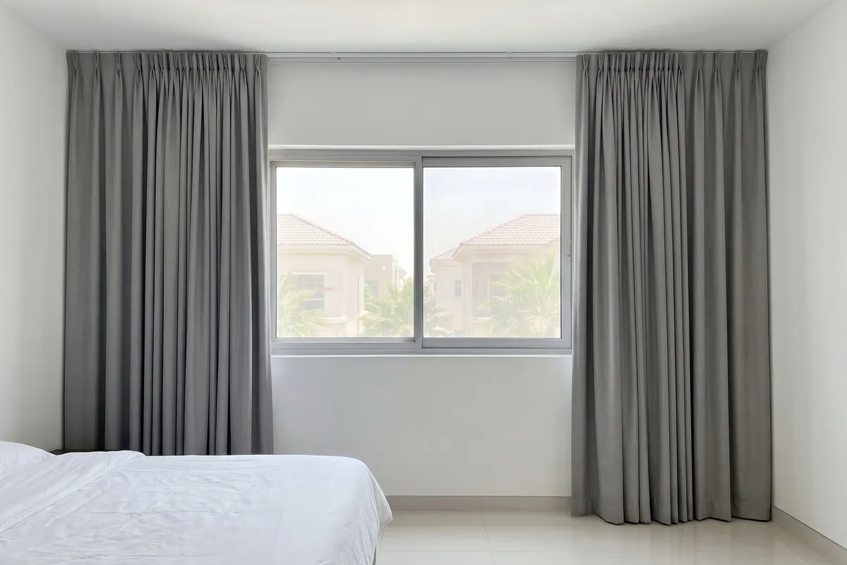 Mid grey curtains drawn open at both sides of a bedroom window on a track fixed tight to the gypsum ceiling, villas outside