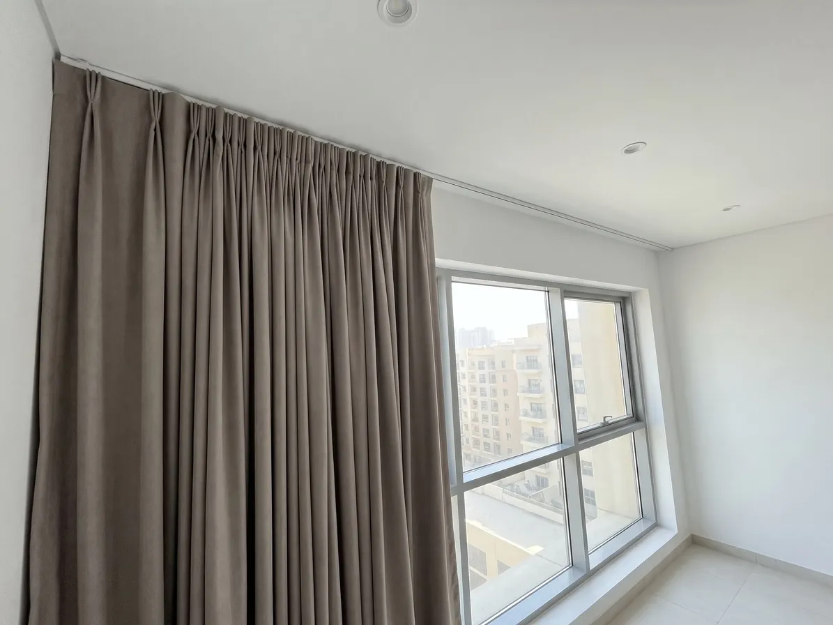 Taupe pinch pleat curtain hanging from a white ceiling track, seen from below at an angle, a window with apartment blocks at the right