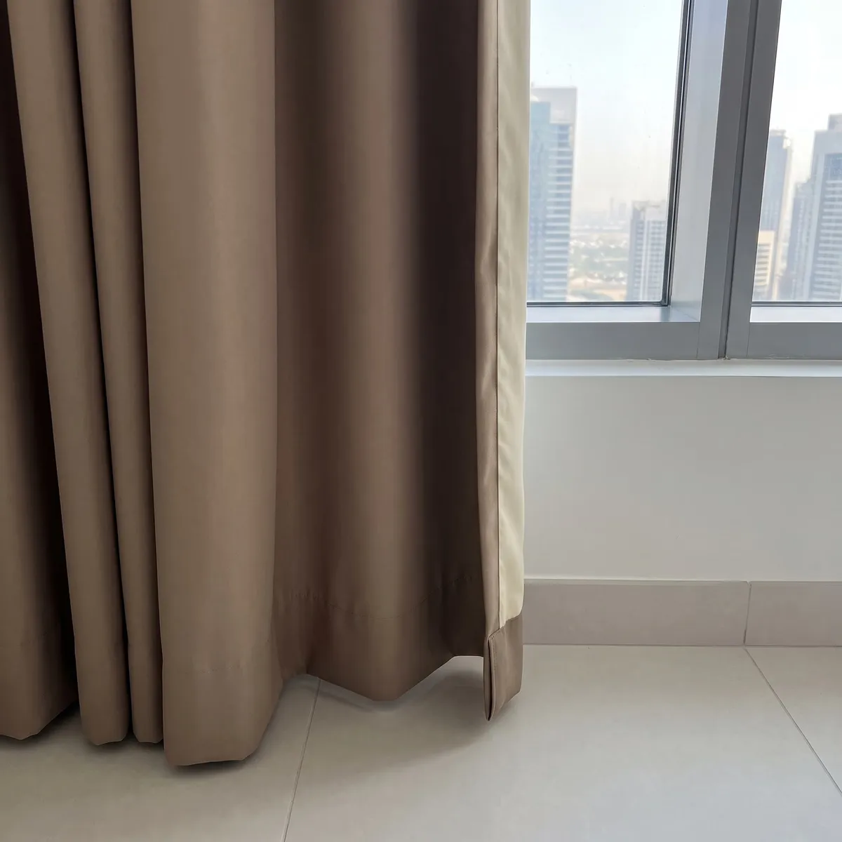 Lower part of a taupe lined curtain, its deep level hem and straight leading edge with the cream lining showing, the floor below