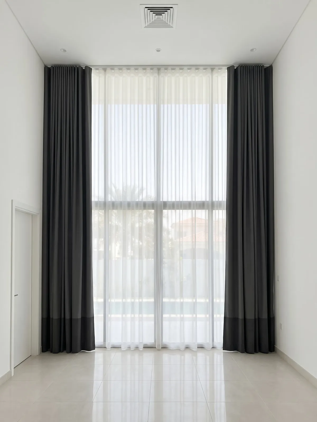 Charcoal lined curtains open at both sides of double-height villa glass, a white sheer closed across it, palms soft behind