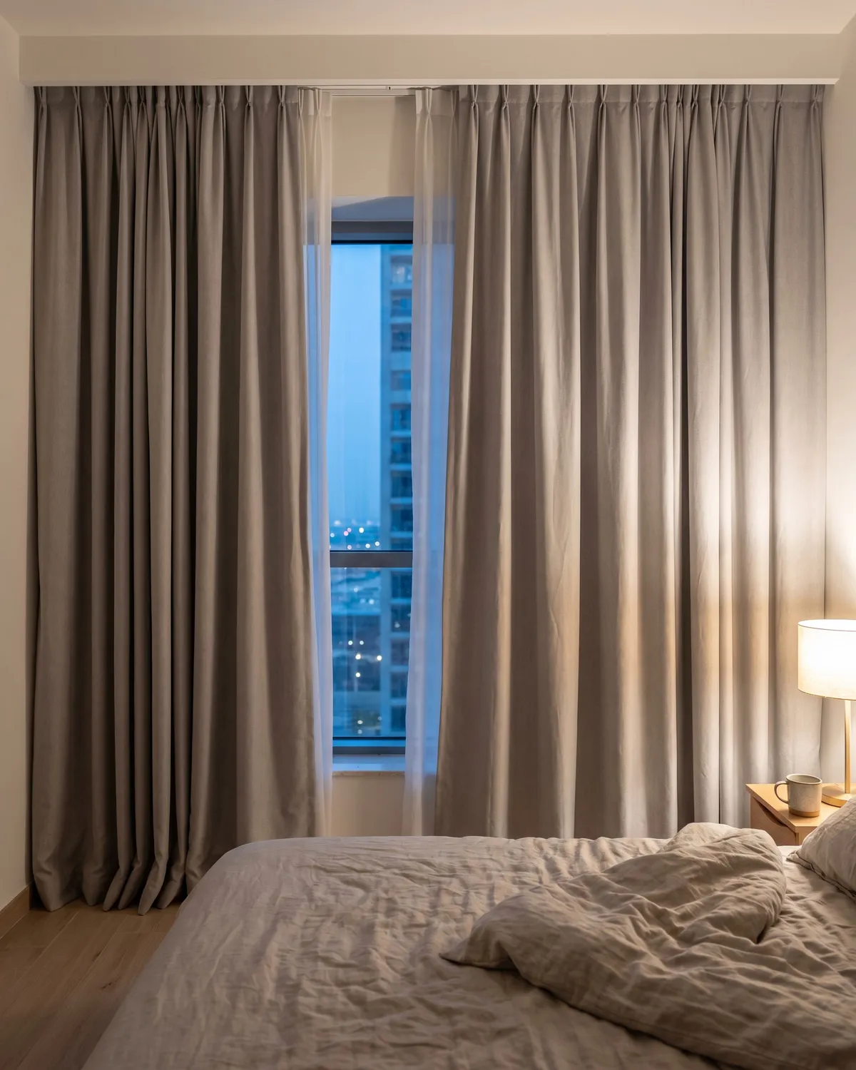 Grey curtains on a ceiling track in a bedroom at dusk, partly open to the city, a bedside lamp on