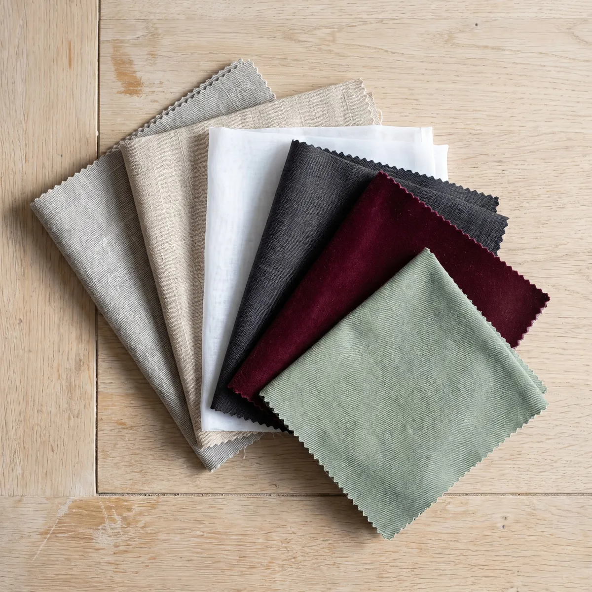 Six curtain fabric swatches fanned out on an oak table: grey, sand, white, charcoal, claret and sage